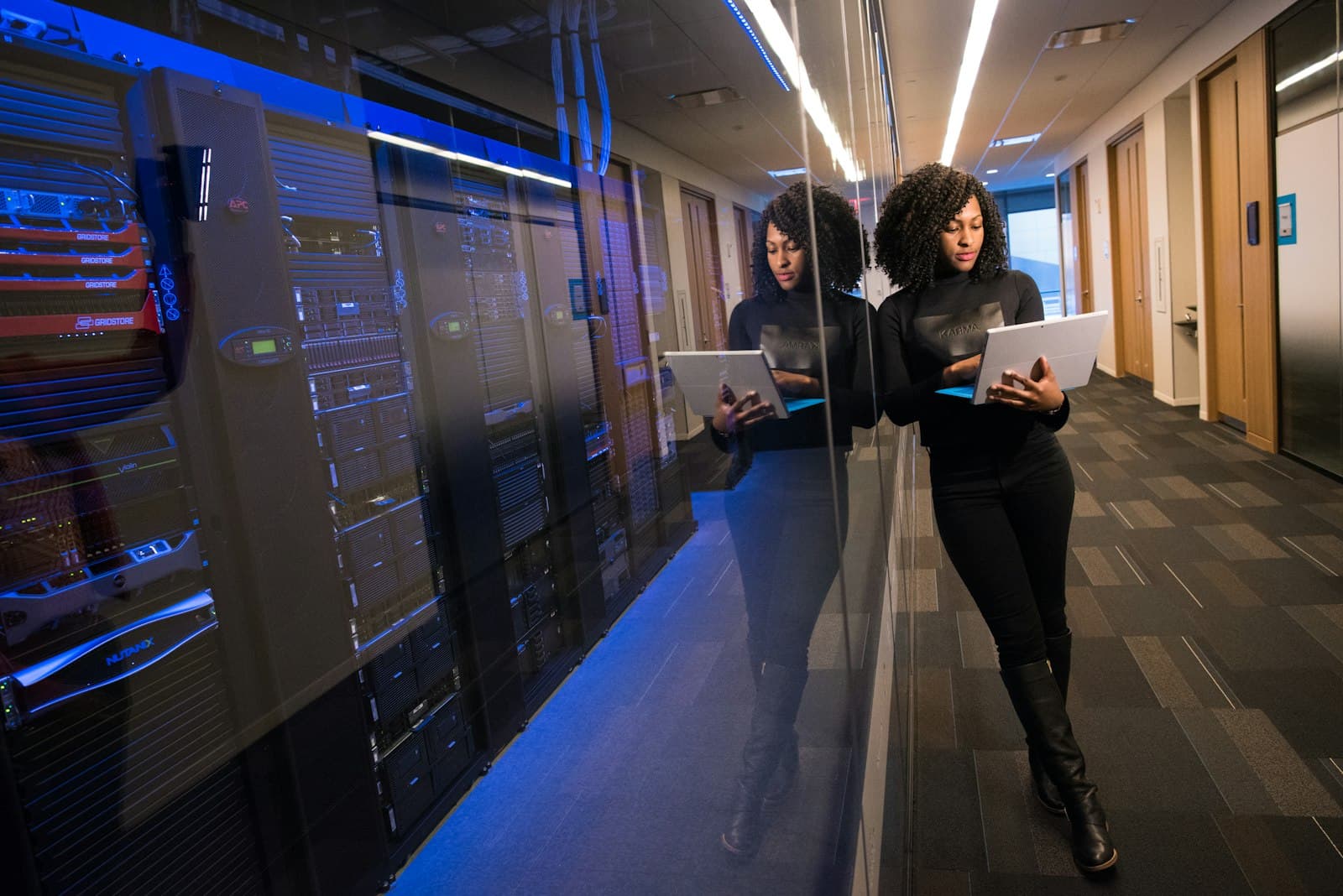 A woman reviewing a tablet, reflected against a dark server rack corridor.